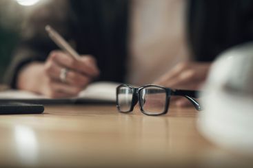 Hands, writing and notebook with glasses in office with...