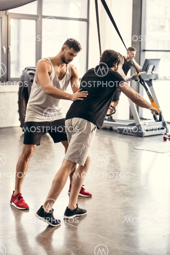 side view of trainer helping sportsman exercising with suspension straps in gym 
