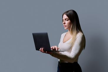 Profile view, young woman using laptop on grey studio...