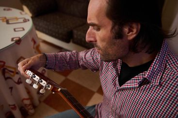 Man tuning a guitar indoors wearing a checkered shirt,...