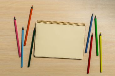 Colorful pencils surround a blank sketchpad on a wooden...
