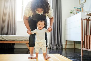 Mother, baby and learning to walk holding hands in home,...