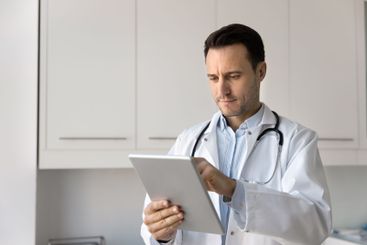 Serious male practitioner doctor using online service on...