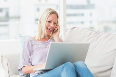 Woman talking on phone while using laptop on sofa
