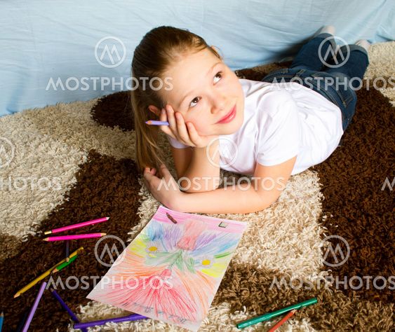 The girl lies on a carpet and thoughtfully draws color pencils a picture