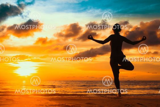 Silhouette young woman practicing yoga on the beach at sunset.