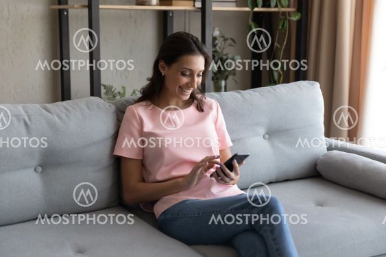 Smiling Arabian woman using smartphone, sitting on couch...