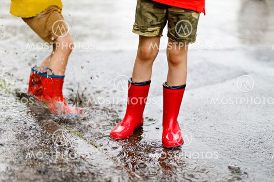 Two children wearing red rain boots jumping into a puddle.