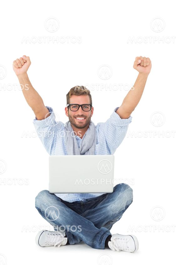 Handsome young man cheering at camera with laptop sitting on floor