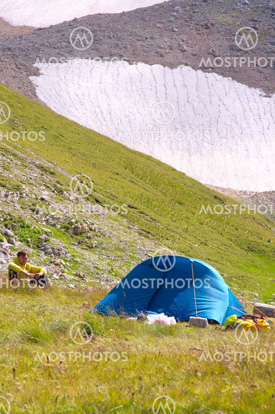 Camping Tent in Mountains with Man Hiker sitting on grass and Glacier Hillside on Background