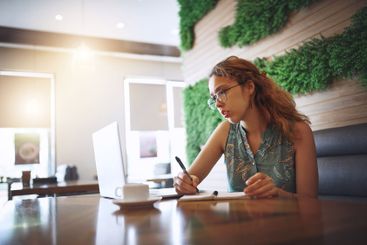 Woman, writing and notebook in coffee shop with remote...