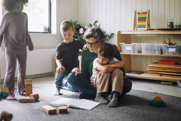 Group of kids sitting with female teacher reading book in...