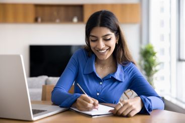 Woman smiles, writes in notebook seated at desk with laptop