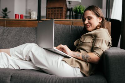 Side view of smiling woman using laptop on sofa in...