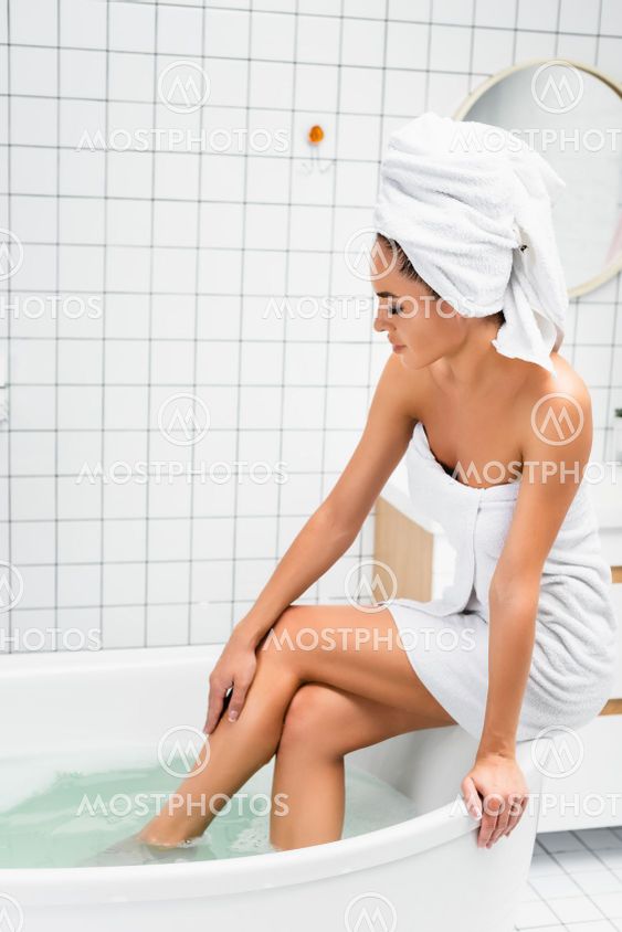 Woman in towels touching leg while sitting on bath with...