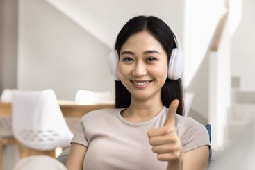 Asian smiling woman wearing headphones, giving thumbs-up...