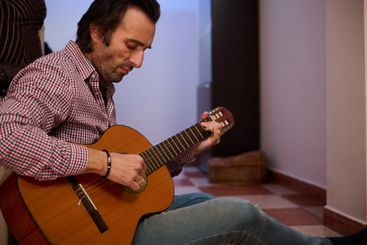 Man playing acoustic guitar while sitting indoors and...