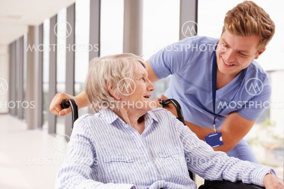 Nurse Pushing Senior Patient In Wheelchair Along Corridor