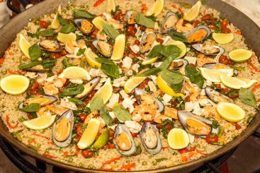 Seafood paella is cooked in a large metal pan. A...