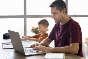 Man working on laptop, his son studying seated at table