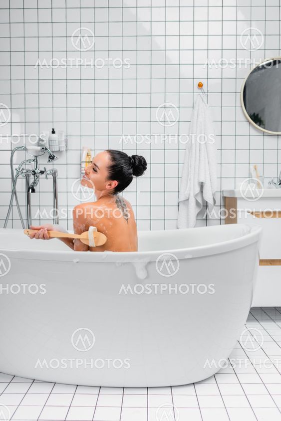 Cheerful tattooed woman cleaning body with brush while...