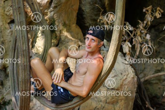 shirtless young man relaxing and leaning against tree or a liana in front of cave