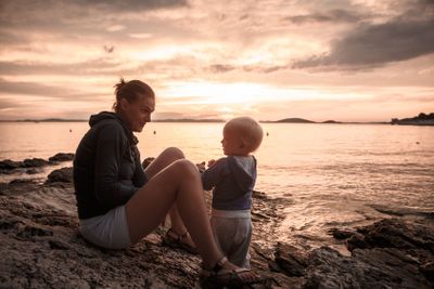 Mother and son sitting on a rocky beach, talking and...