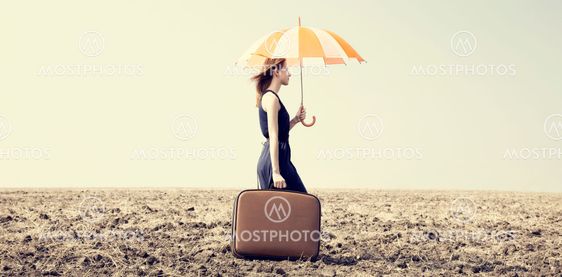 Redhead girl with umbrella and suitcase at windy grass meadow.
