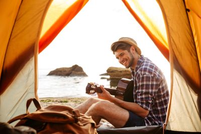 Smiling young man sitting near touristic tent and playing...