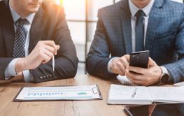 Two men in a suit are sitting at a desk with graphs...