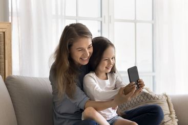 Smiling young mom little daughter cuddle on couch using...