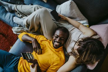 Cheerful man lying on sofa with girlfriend sitting in...