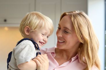 Loving Mother Cuddling Young Son At Home In Kitchen...