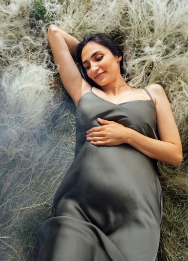 Young pregnant woman in green satin dress lies on grass...