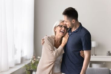 Happy older mom stand by window hugging beloved adult son