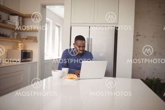 Smiling man using laptop while having breakfast in kitchen
