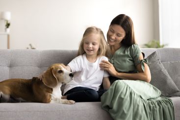 Little girl her mother sits on sofa, gently petting dog