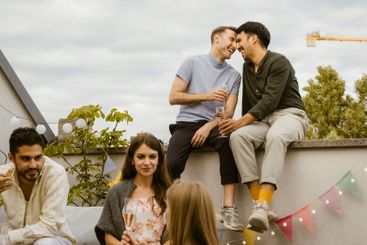 Happy gay couple sitting on wall with friends talking...