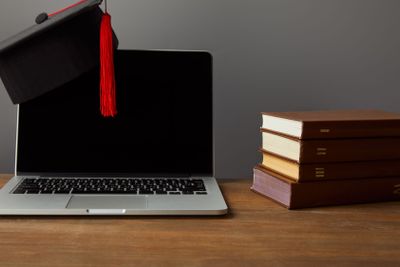 Laptop with blank screen, books and academic cap with red...