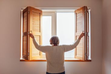An elderly woman in a sweater opens the wooden shutters....