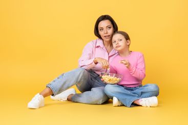 Mom and daughter enjoy popcorn together against a yellow...