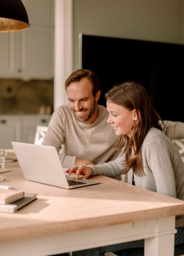 Smiling father sitting by daughter using laptop for...