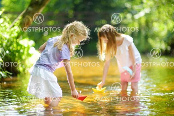 Two little sisters playing with paper boats by a river on...