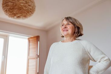 An elderly woman in a knitted sweater looks out the...