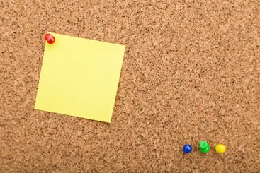 Yellow sticky note on corkboard with colored push pins...
