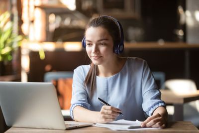 Focused woman wearing headphones using laptop, writing...