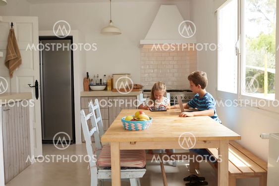 Children At Home Using Digital Devices On Kitchen Table