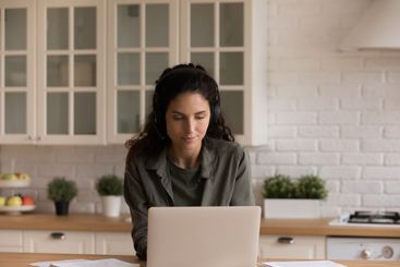 Young woman in headset work on laptop from home kitchen