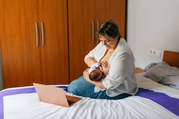 Mother breastfeeding newborn baby while working from...