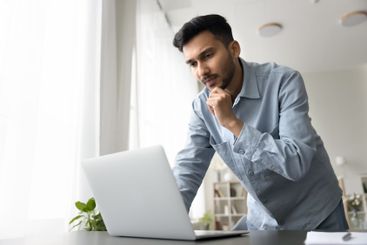 Pensive millennial male stand by desk look on notebook...